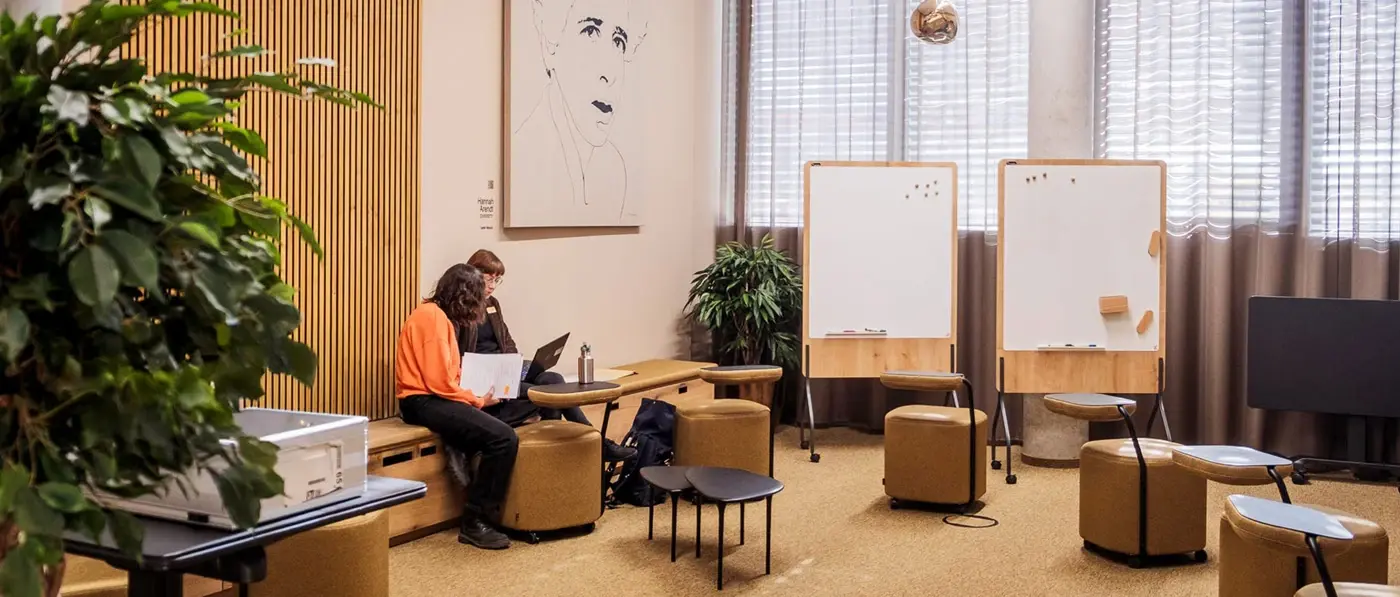 Konferenzraum mit mehreren runden Hockern, zwei Whiteboards, hängenden Kugelleuchten und zwei Personen an einem Laptop