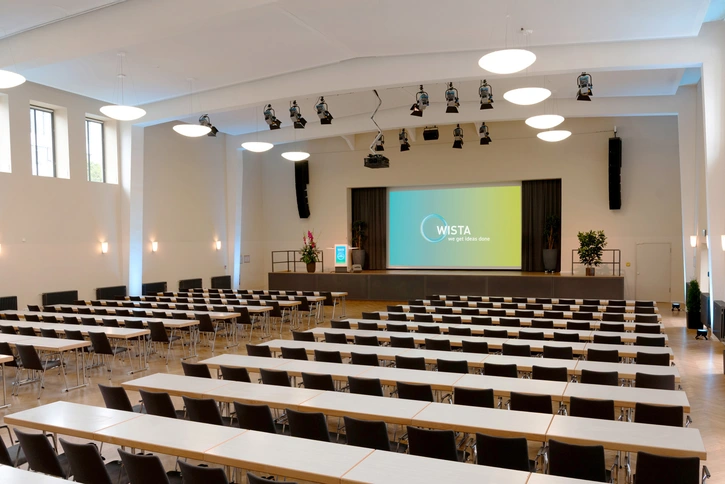 Bunsen-Saal mit Reihen von Tischen und Stühlen sowie Bühne mit Leinwand