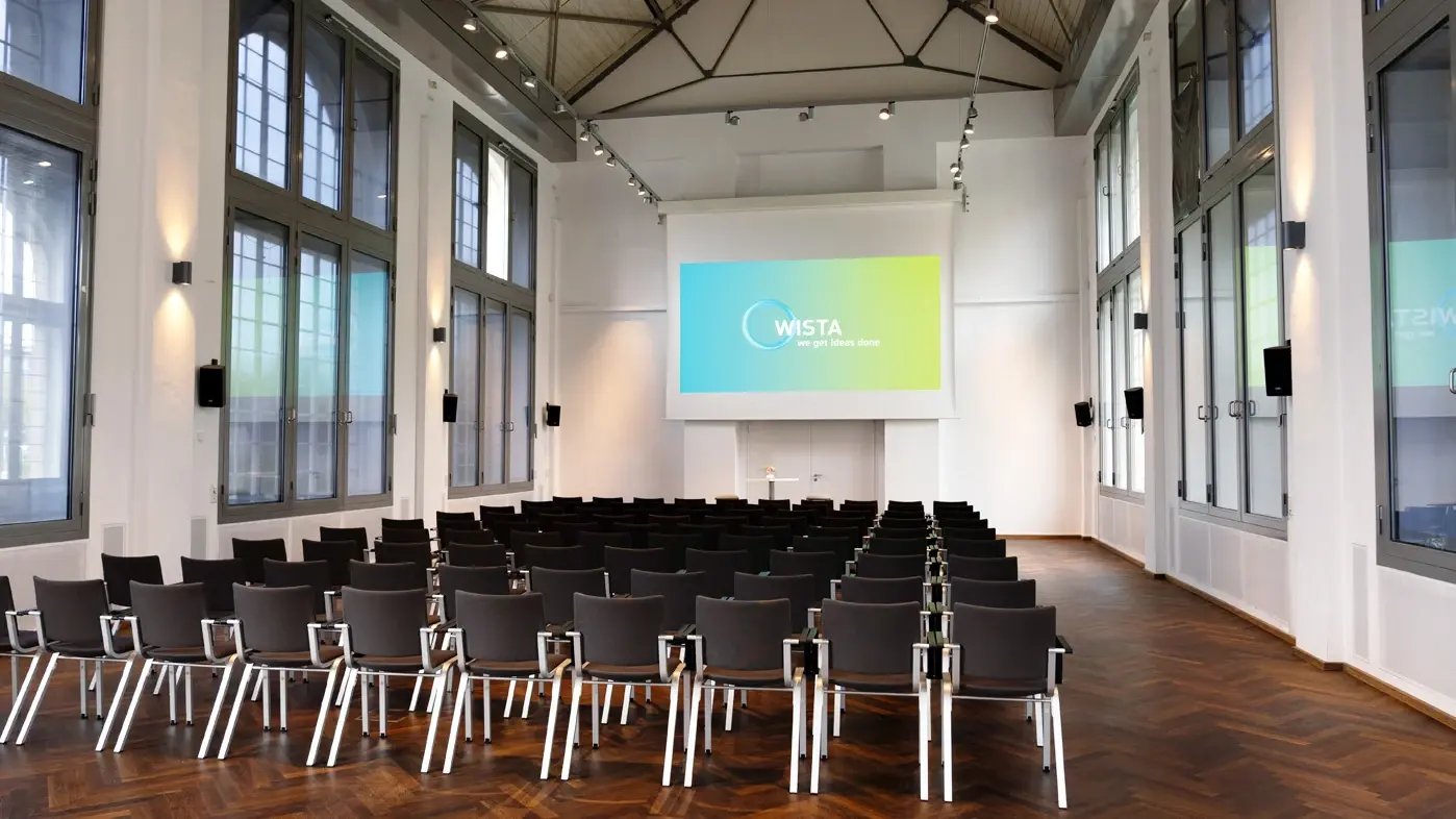Hans-Grade-Saal mit Reihen von Stühlen vor einer Leinwand und hohen Fenstern an den Seiten
